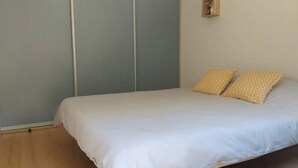 Una cama individual con ropa blanca y dos almohadas estampadas, una mesita de noche pequeña y una lámpara fijada a la pared.