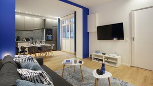 Una sala de estar moderna con un sofá gris, una mesa de centro y un comedor con sillas y una mesa. Un televisor de pantalla plana está montado en la pared, y hay un mueble blanco debajo.