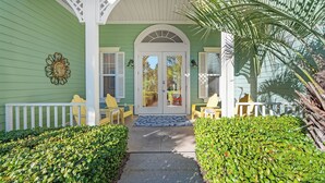 A green house with a white porch, a decorative wall piece, and a palm tree. - Casa Del Mar - Beautiful New Rental (Gulf Shores)