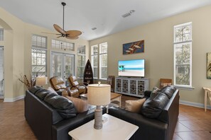 A spacious living room with a ceiling fan, a television, and a view of the ocean through large windows. - Casa Del Mar - Beautiful New Rental (Gulf Shores)