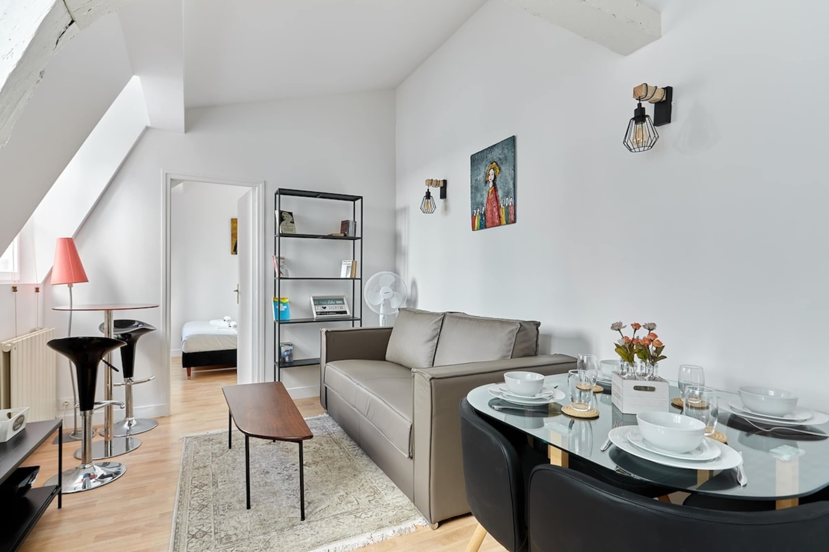 A modern living room with a sofa, a dining table set with bowls and glasses, a bookshelf, and a painting on the wall.