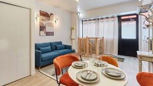 A modern living room with a blue sofa, orange chairs, a dining table set for four, and a chandelier. - Charmant Duplex 1 Chambre Centre de Paris (Paris)