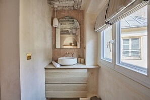 A bathroom with a white sink, wooden drawers, a towel, a mirror, and a window with a view of the outside. - Amazing Apartement Place Vendome/rue de la Paix (Paris)
