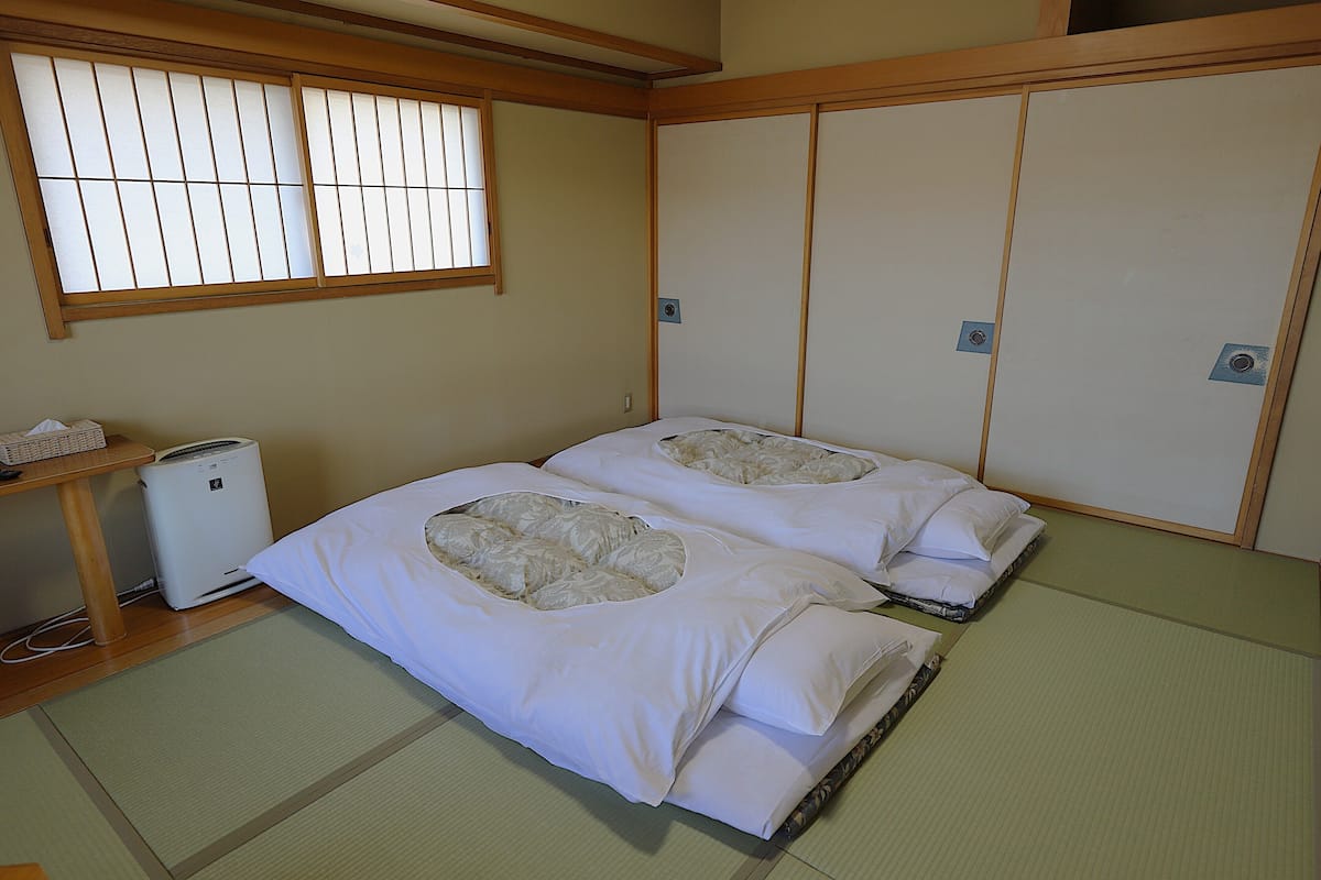 Ein traditioneller japanischer Raum mit Tatami-Bodenbelag, zwei Tatami-Matten, einem Futonbett mit Decke, einem kleinen Heizgerät und einem Fenster mit Holzrahmen.