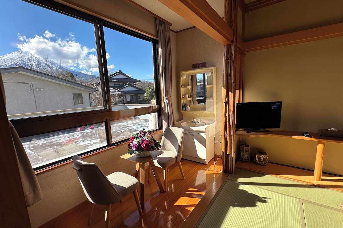 Ein Zimmer mit Holzboden, einem Fernseher, einem Waschbecken, einem Sessel, einem Tisch mit Blumenvase und Blick auf ein Gebäude mit Bergen im Hintergrund.