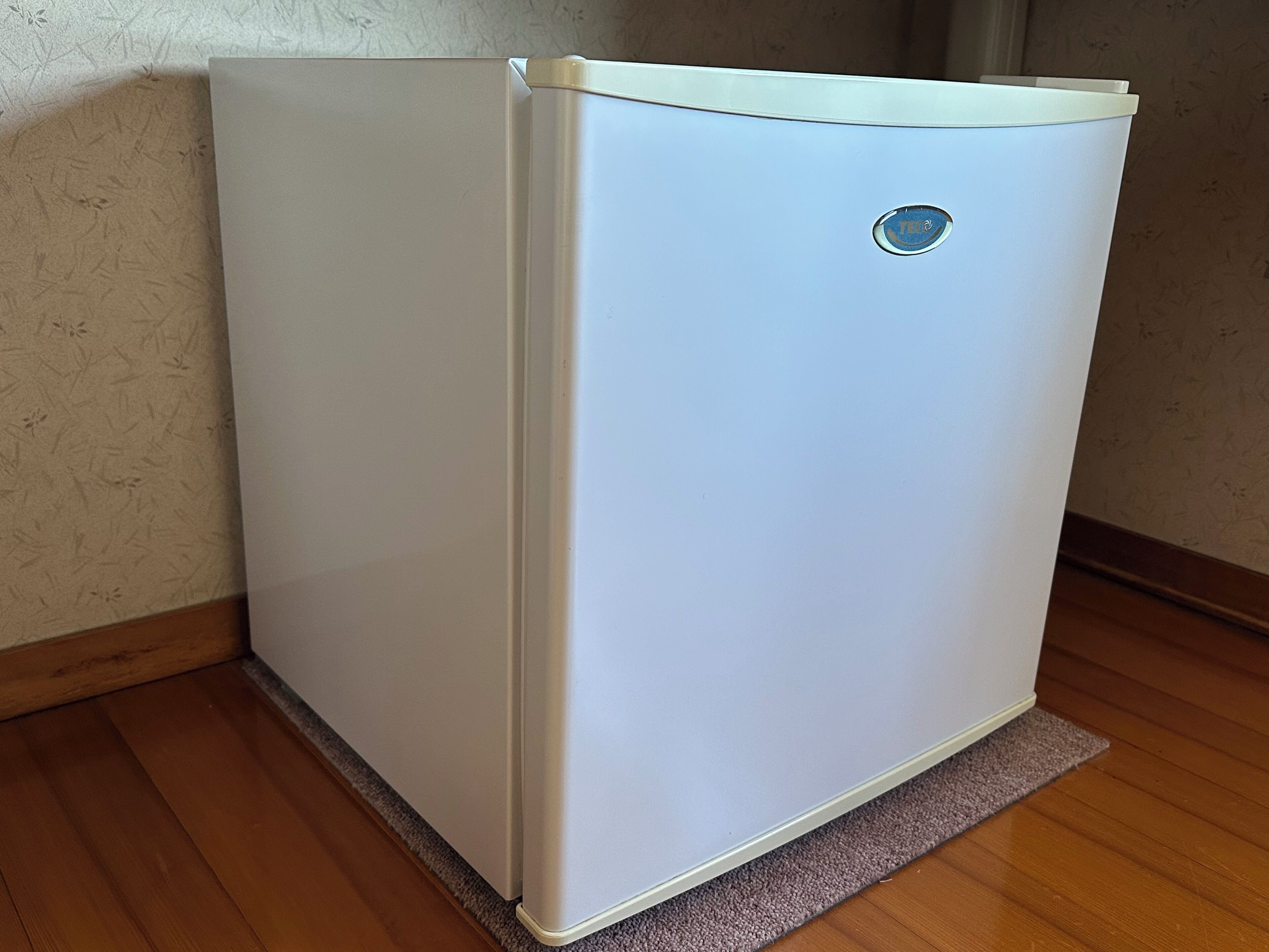 A white refrigerator with a blue logo, positioned against a wall with a textured wallpaper and wooden flooring.