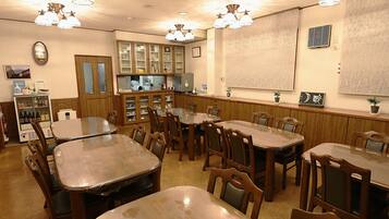 Un comedor con varias mesas y sillas de madera, una zona de cocina con armarios y electrodomésticos, y una pantalla de proyector fijada a la pared.