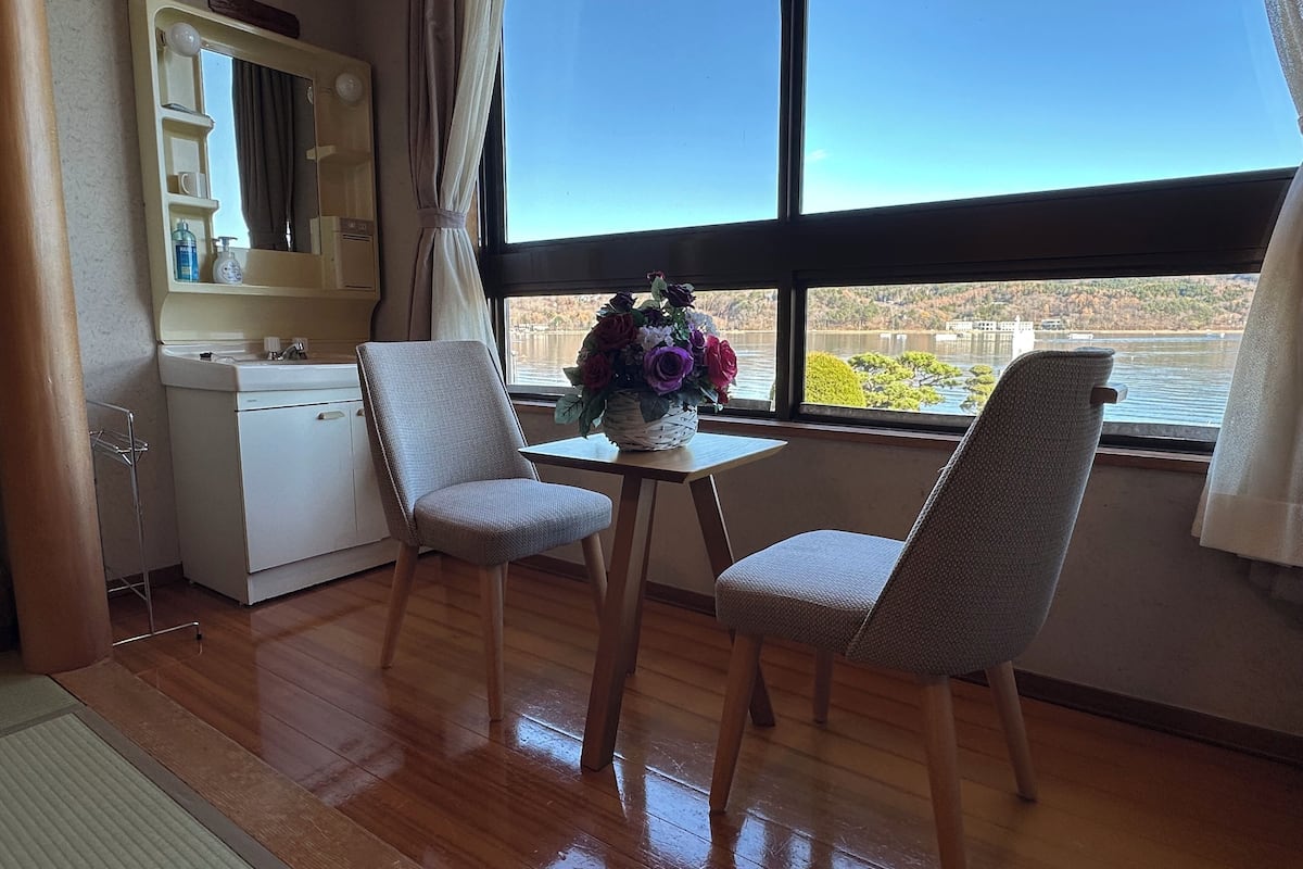 Ein Zimmer mit zwei Sesseln, einem kleinen Tisch mit Blumenvase, einem Fenster mit Blick auf ein Gewässer und einem Spiegel an der Wand.