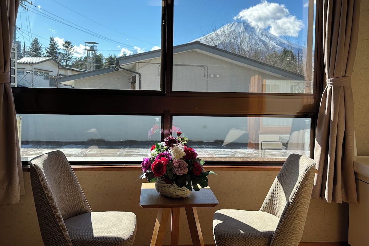 Ein Zimmer mit zwei Stühlen, einem kleinen Tisch mit Blumenvase und einem großen Fenster mit Blick auf Berge und Häuser.