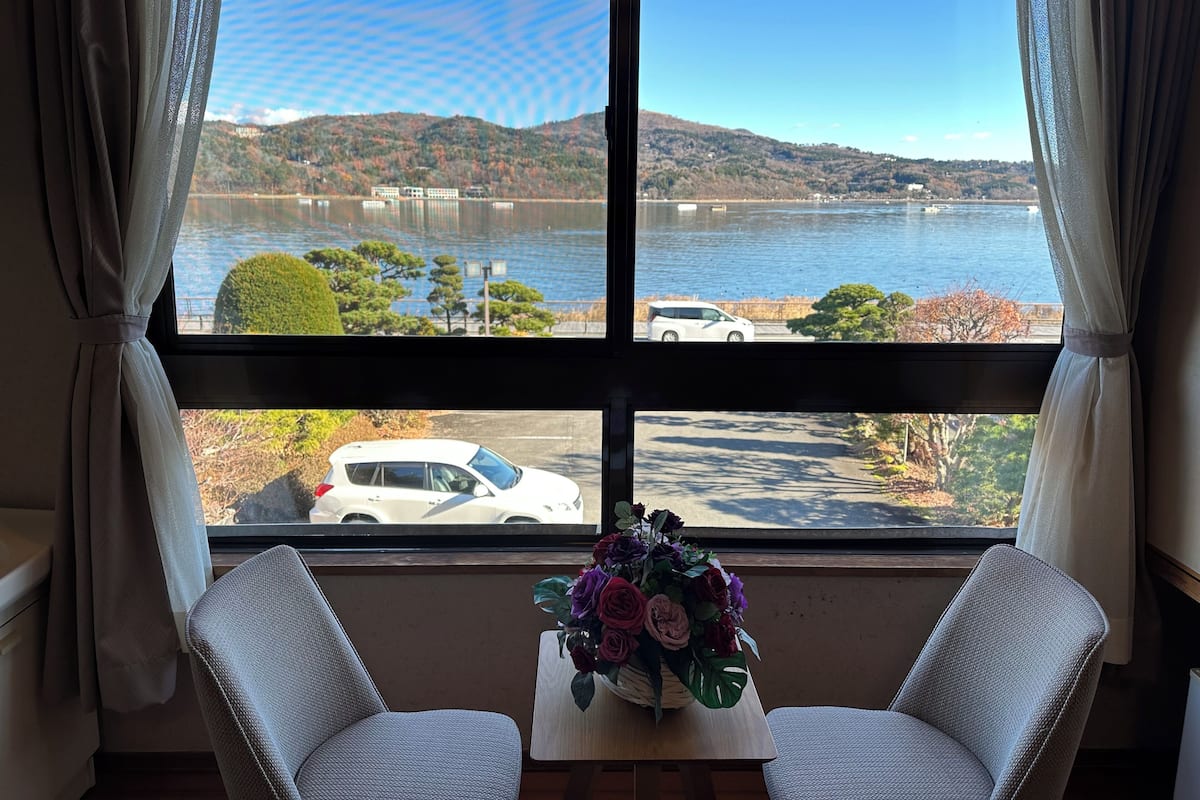 Ein Zimmer mit großem Fenster mit Blick auf einen See, ein weißes Auto ist vor dem Haus geparkt und ein Tisch mit einer Blumenkomposition.