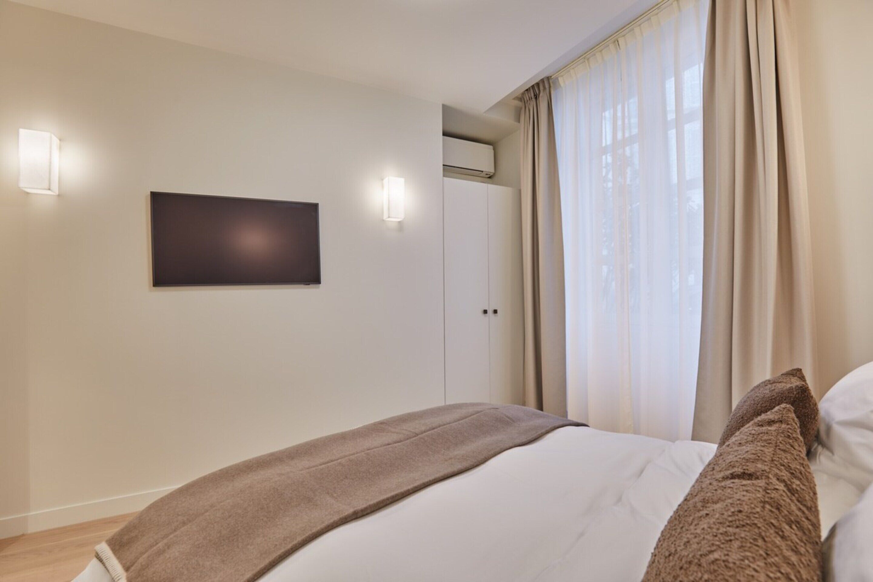 A bedroom with a bed, a flat-screen TV mounted on the wall, and a window with curtains.