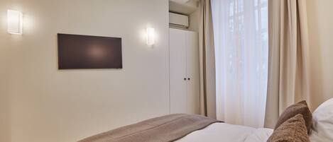 A bedroom with a bed, a flat-screen TV mounted on the wall, and a window with curtains.