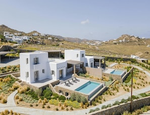 A modern villa with a pool and outdoor seating, surrounded by landscaped gardens and set against a hilly backdrop. - Pristine Villa With Swimming Pool (Mykonos)