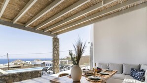 A terrace with a wooden table set for dining, a stone pillar, and a view of the sea. - Pristine Villa With Swimming Pool (Mykonos)