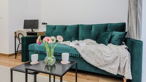 Un canapé vert surmonté d’un plaid blanc, une petite table avec deux tasses blanches et un vase rempli de fleurs, et un téléviseur posé sur un meuble en bois.