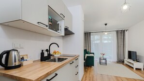 Une cuisine moderne équipée d’un four à micro-ondes, d’une bouilloire et d’un panier à fruits. On y trouve également un coin repas avec une chaise verte et un téléviseur. La pièce est dotée d’un plancher en bois et de murs blancs.