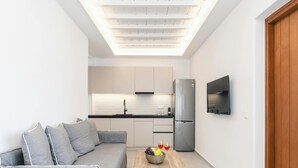 Ein kompakter Wohnbereich mit einer Couch, einem Holztisch mit Früchten, einer Küchenzeile inklusive Kühlschrank und einem an der Wand montierten Fernseher.