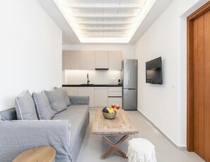 Ein kompakter Wohnbereich mit einer Couch, einem Holztisch mit Früchten, einer Küchenzeile inklusive Kühlschrank und einem an der Wand montierten Fernseher.