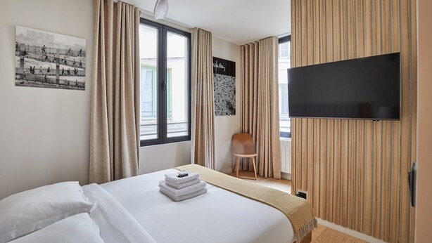 A hotel room with a bed, a ceiling fan, a TV mounted on the wall, and a window with curtains.
