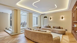 Ein modernes Wohnzimmer mit einem großen beigen Sofa, einem Couchtisch und einer Stehlampe. Eine Treppe führt zu einer oberen Ebene, und ein Bücherregal zeigt Bücher und Dekorationsobjekte.