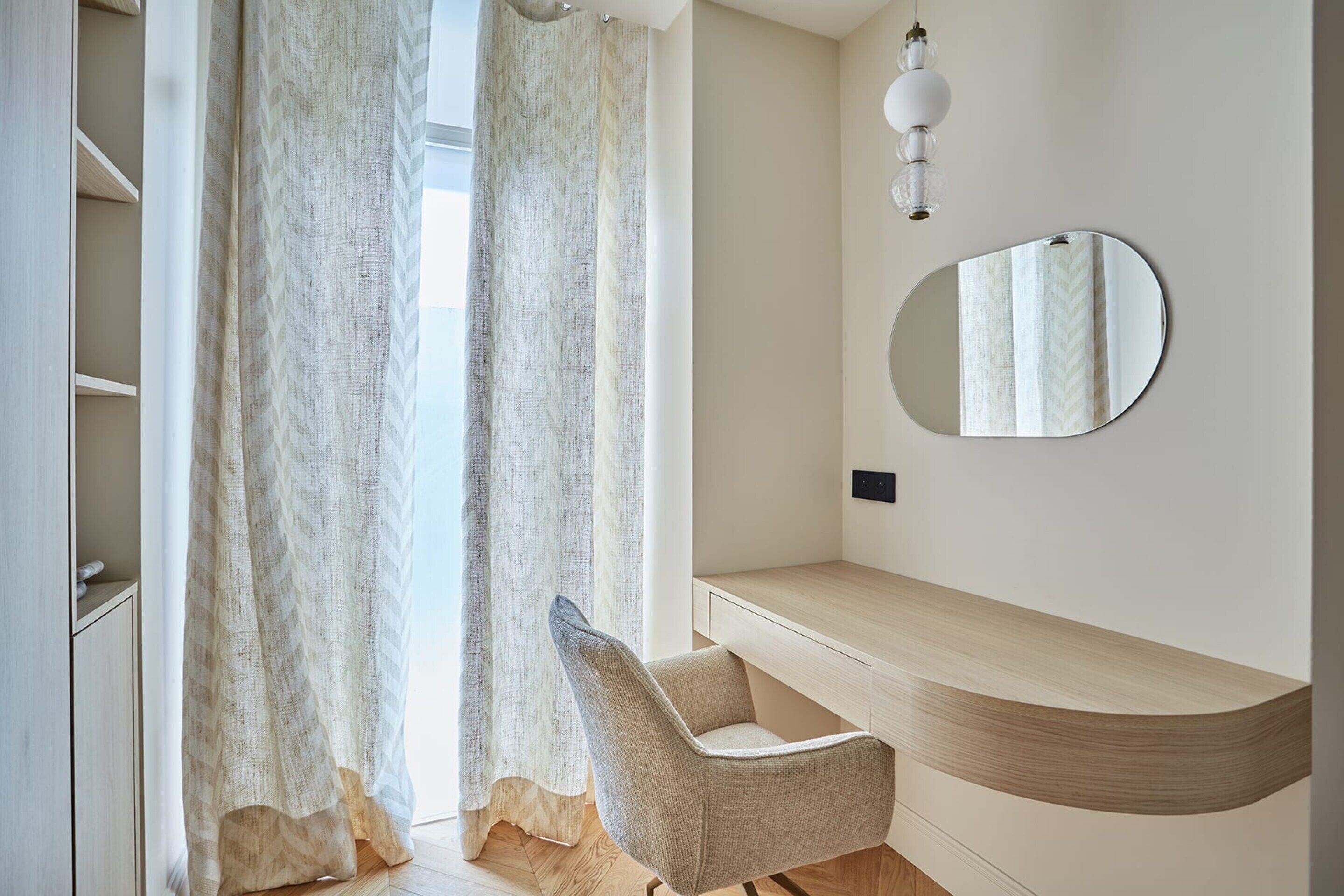 Un dormitorio moderno con una mesita de noche de madera curvada, una silla y un espejo grande en la pared.