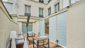 Ein Innenhof mit Holzboden, einer Sitzecke mit weißen Kissen und einer Glasschiebetür, die auf einen Balkon führt.