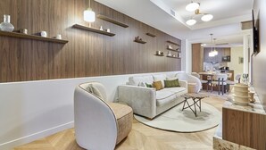 Un salon moderne avec un canapé, un fauteuil et une table basse, des étagères en bois et, au fond, un coin repas.
