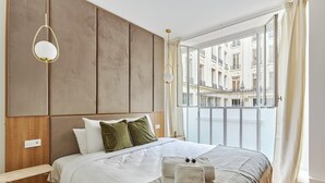 Une chambre dotée d’un grand lit, d’une tête de lit en bois, de tables de chevet et offrant une vue sur le paysage urbain grâce à un balcon.