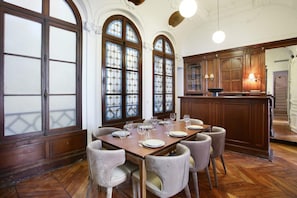 A dining room with a wooden table set for six, flanked by arched windows with stained glass, a wooden cabinet, and a kitchen area in the background. - Magnificent Apartment With AC - Louvre Museum (Paris)