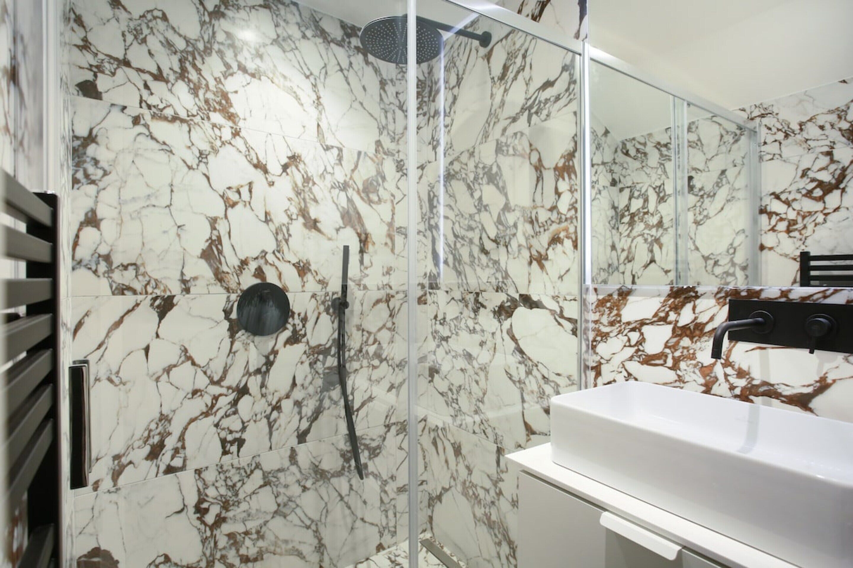 A bathroom with a marble wall, a glass shower enclosure, and a white sink.