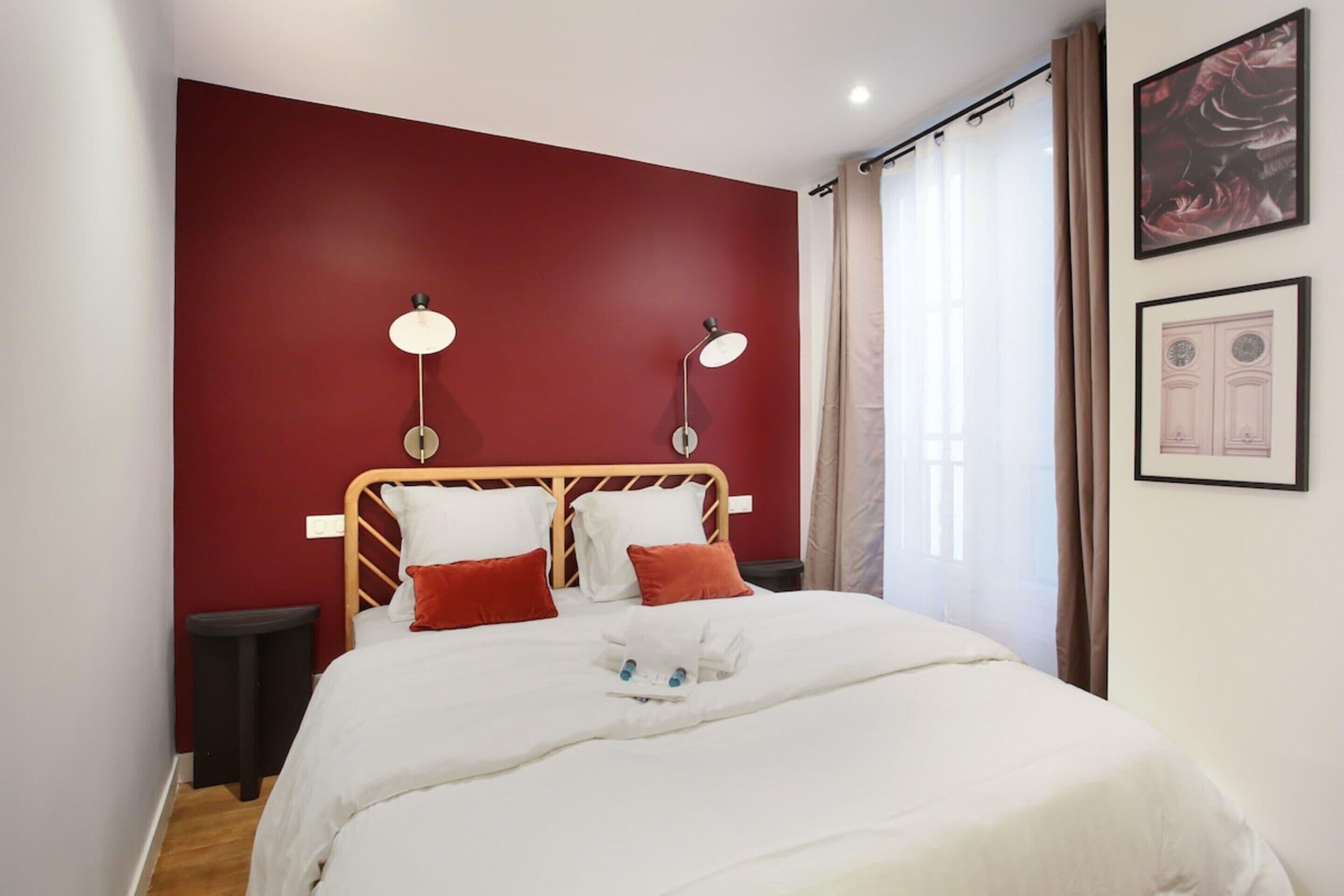 A neatly made bed with white linens and red pillows, two bedside lamps, a small bedside table, and a framed picture on the wall.