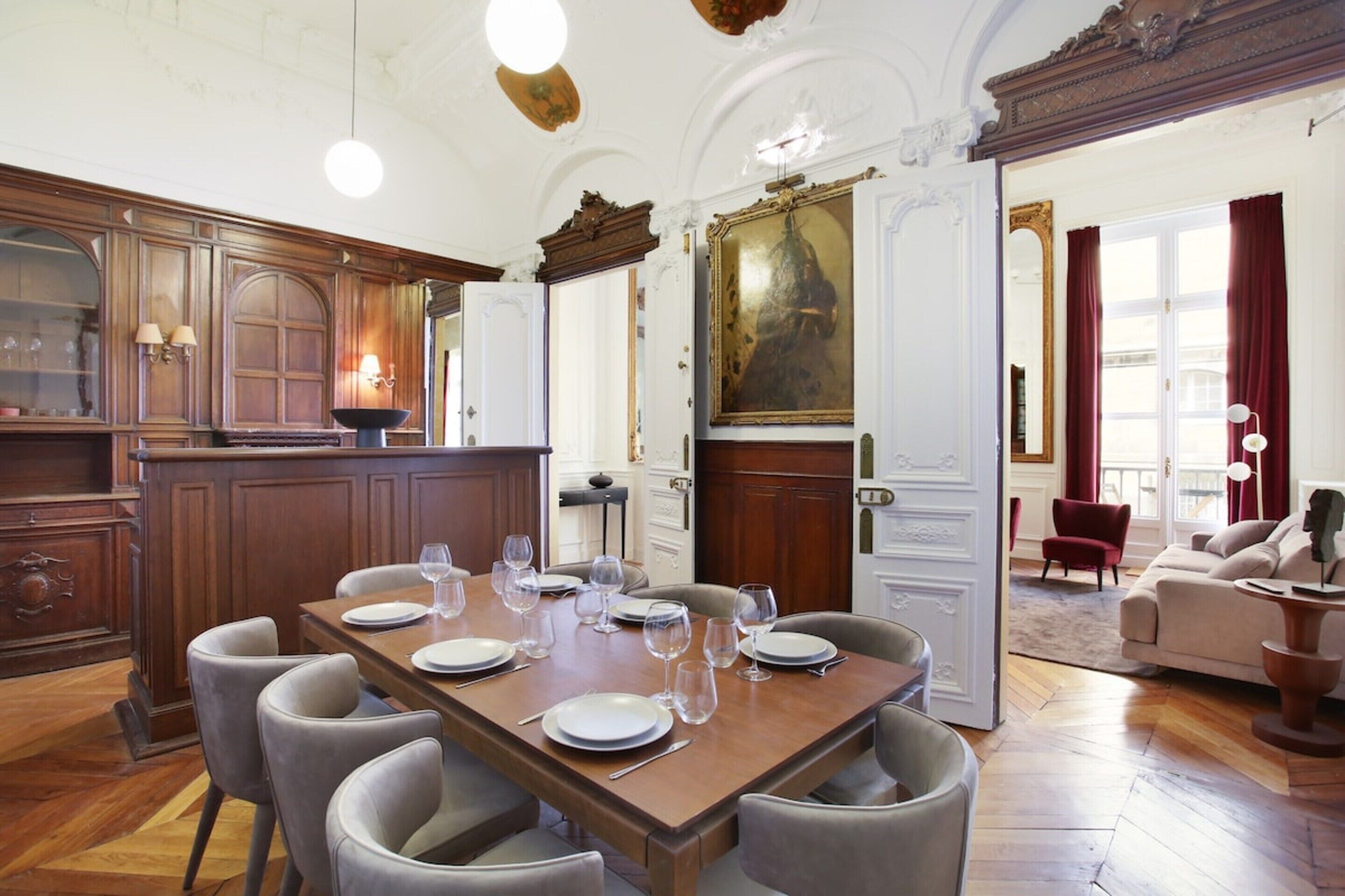 Un comedor con una mesa de madera dispuesta para seis personas, rodeada de sillas. Hay un cuadro grande sobre la puerta y un candelabro colgando del techo.