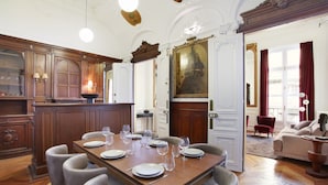 Un comedor con una mesa de madera dispuesta para seis personas, rodeada de sillas. Hay un cuadro grande sobre la puerta y un candelabro colgando del techo.
