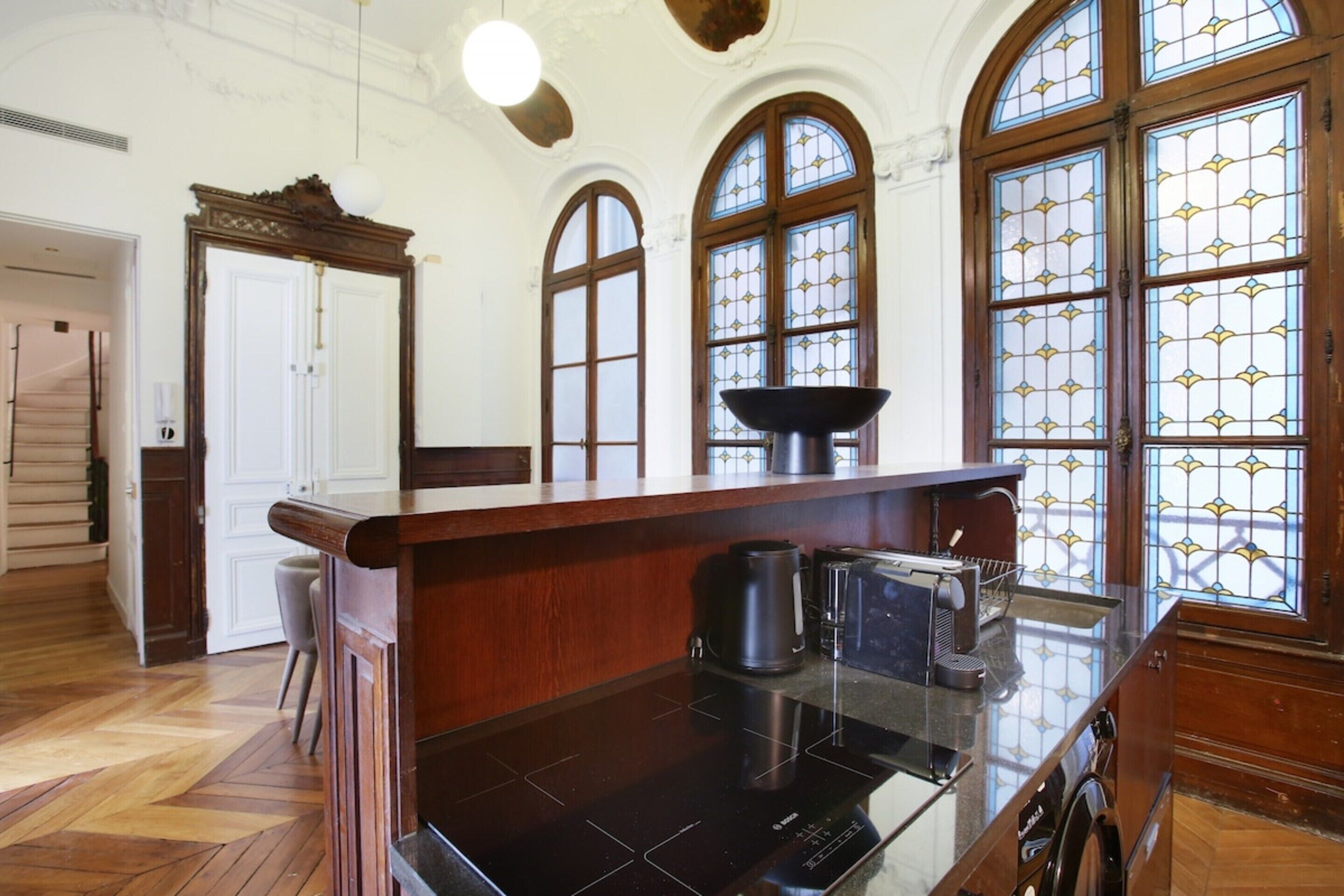 A kitchen with a wooden countertop, a coffee maker, and a toaster. There are arched windows with stained glass.