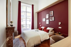A bedroom with a bed, a wooden floor, a marble fireplace, a window with curtains, and a red accent wall. - Magnificent Apartment With AC - Louvre Museum (Paris)
