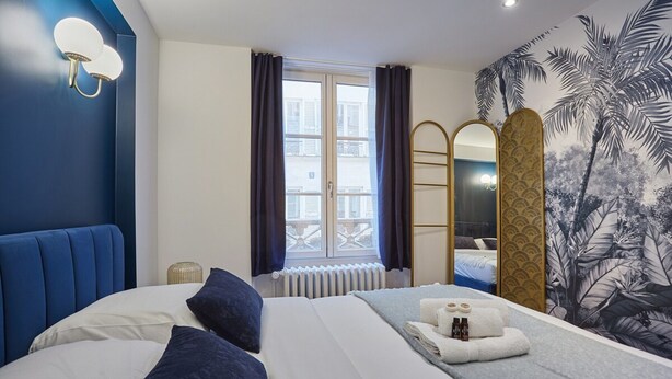 A hotel room with a large bed, a window with curtains, a radiator, a towel and toiletries on the bed, a mirror, and a wall with a tropical mural. - Sublim & New Aparment Saint Germain des Pres (Paris)