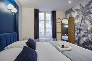 Une chambre d’hôtel avec un grand lit, une fenêtre avec des rideaux, un radiateur, une serviette et des articles de toilette posés sur le lit, un miroir et un mur orné d’une fresque tropicale.