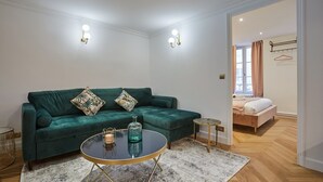A living room with a green sofa, a round coffee table, and a small side table. There is a bed with a pink blanket in the background. - Sublim & New Aparment Saint Germain des Pres (Paris)