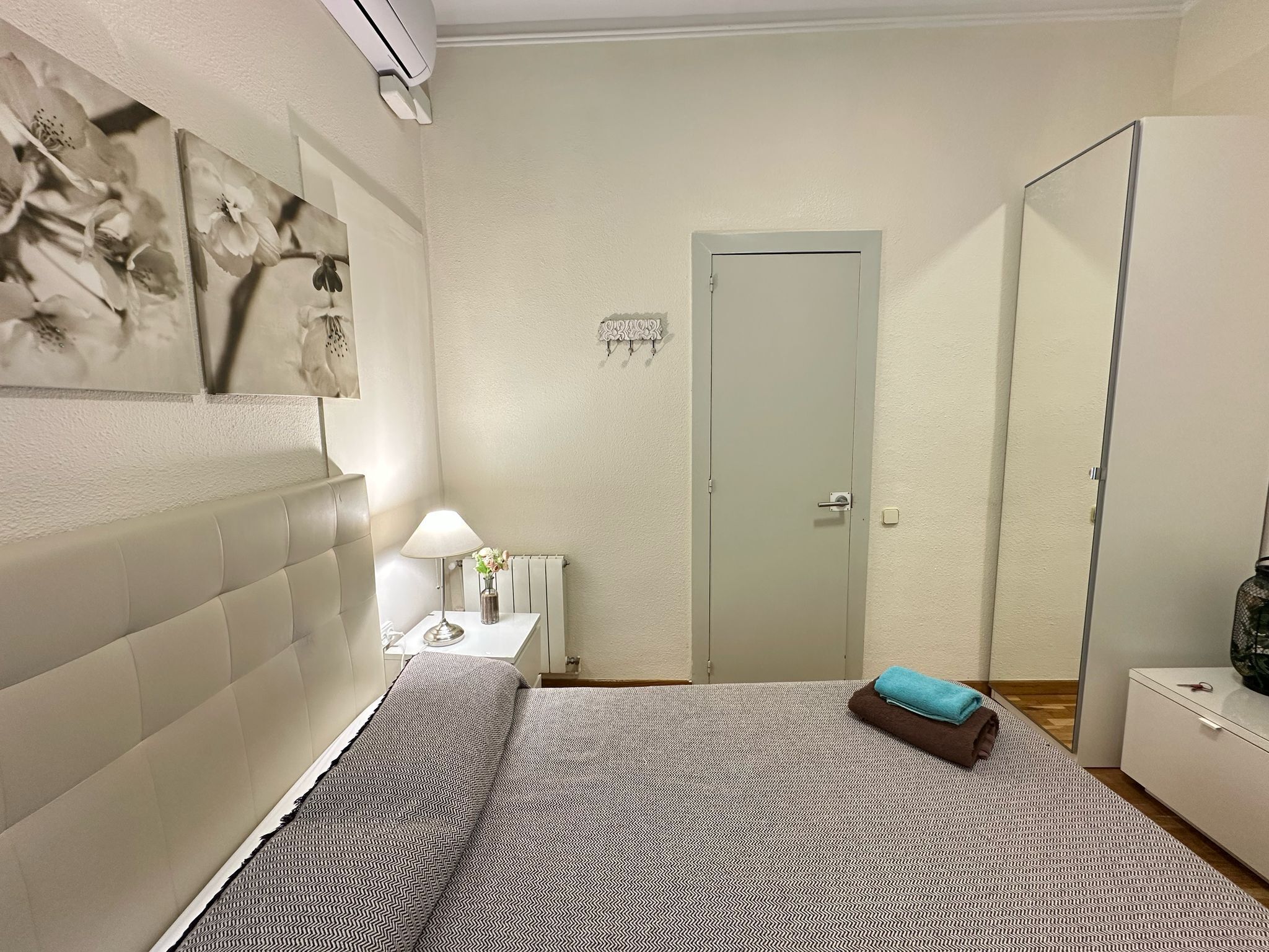 A bedroom with a bed, a bedside table with a lamp, a mirror, and a door.