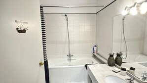 Een badkamer met een witte wastafel, een spiegel en een douche. De vloer is zwart-wit geruit en er staat een bordje met daarop 