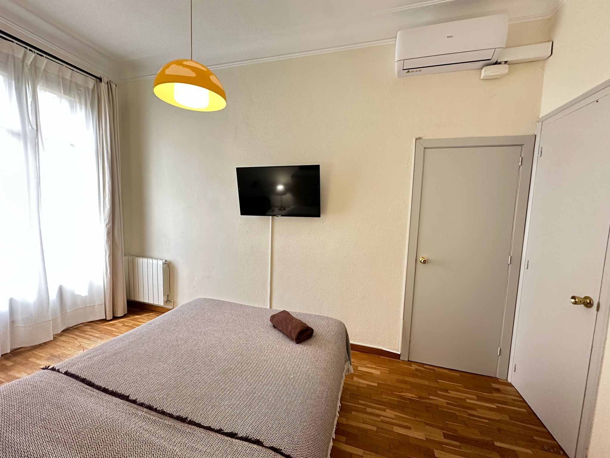A bedroom with a bed, a TV mounted on the wall, a radiator, and an air conditioning unit.