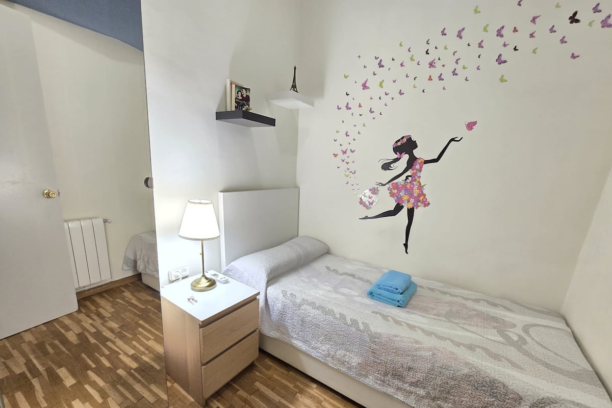 Quarto com cama, mesa de cabeceira com luminária, cômoda, radiador e parede com adesivo de borboleta.