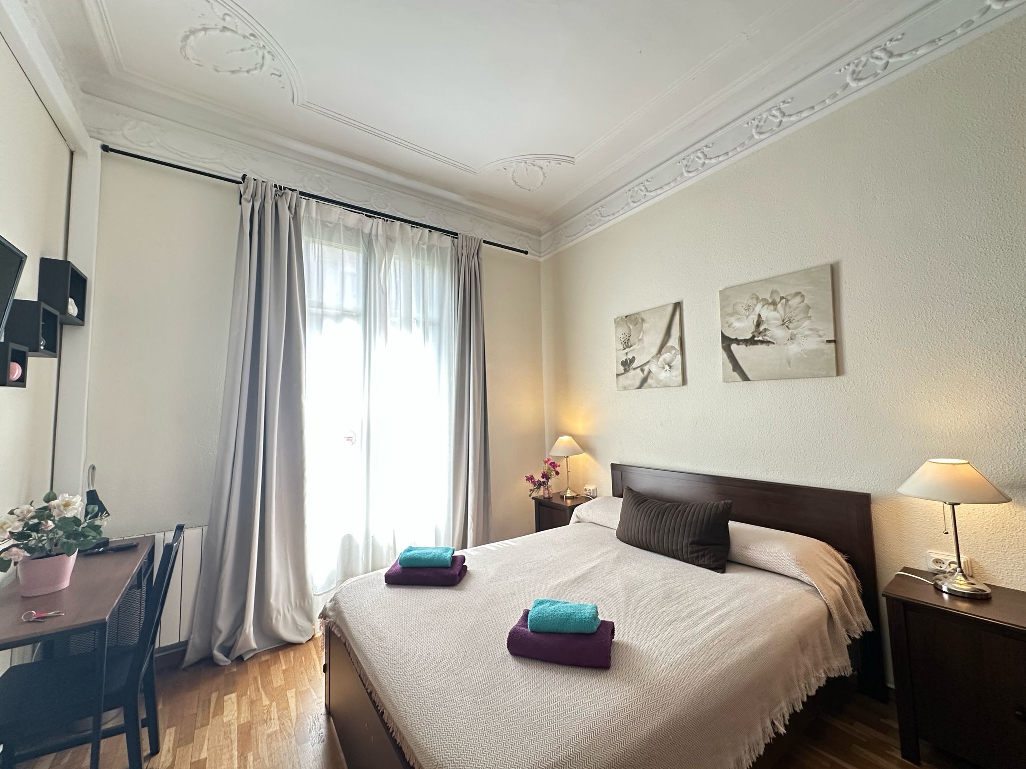 A hotel room with a bed, a desk, a chair, a potted plant, and two wall-mounted lamps.