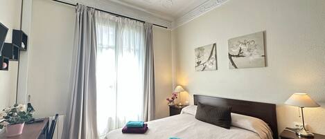 A hotel room with a bed, a desk, a chair, a potted plant, and two wall-mounted lamps.