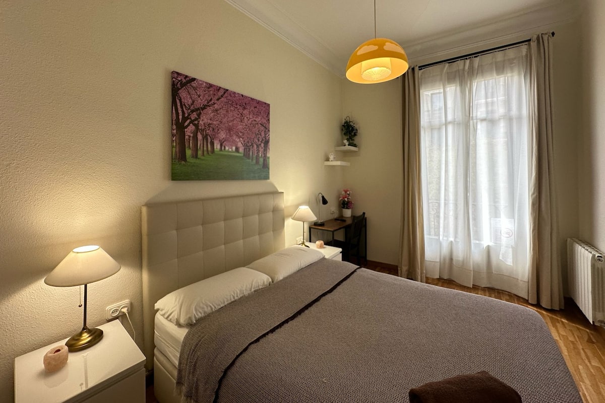 Quarto com cama, abajures, um quadro, janela com cortinas e radiador.