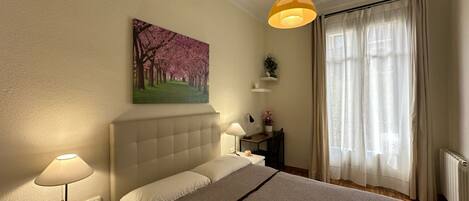 A bedroom with a bed, bedside lamps, a painting, a window with curtains, and a radiator.