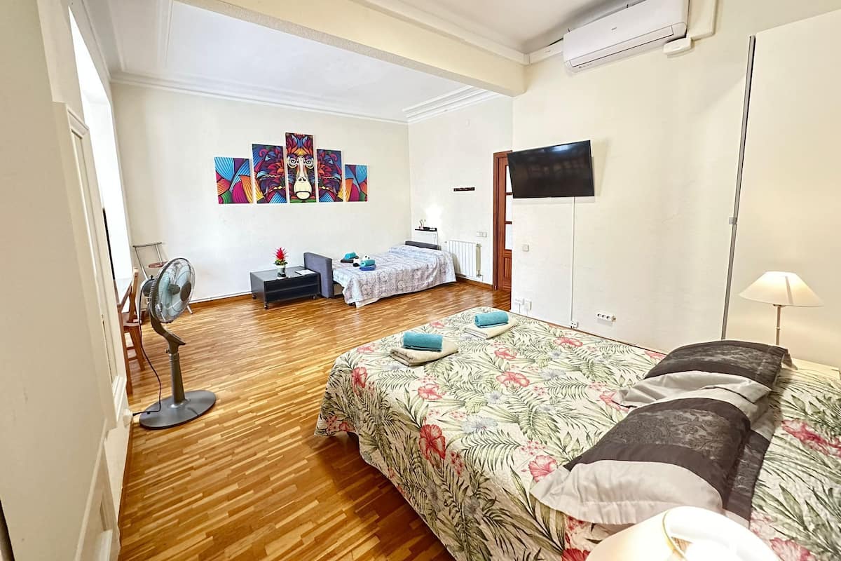 Quarto de hotel com chão de madeira, cama com roupa de cama floral, escrivaninha com cadeira, ventoinha de chão, televisão na parede e janela com vitrais.