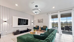A modern living room with a large flat-screen TV, green sofas, a coffee table, and a view of the outdoors through large windows.