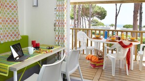 A dining area with a wooden deck, white chairs, a table with a laptop, and a view of trees and the sea. - Bungalow Near Pals Beach & Pool (Pals)