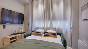 Een moderne slaapkamer met een bed, een flatscreen televisie aan de muur, een nachtkastje met een lamp en dunne gordijnen.